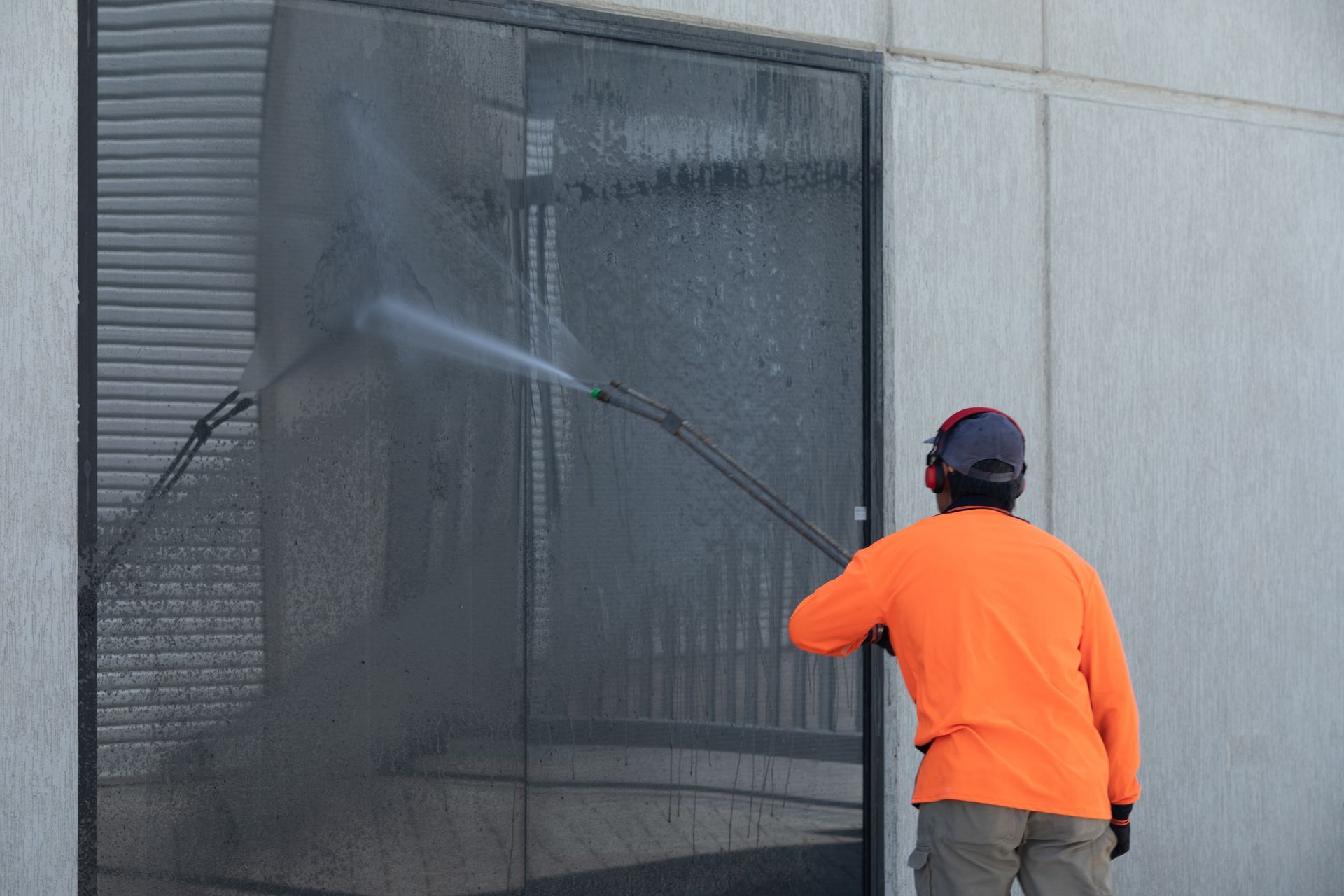 Person in orange jacket pressure-washing a large window on a gray building
