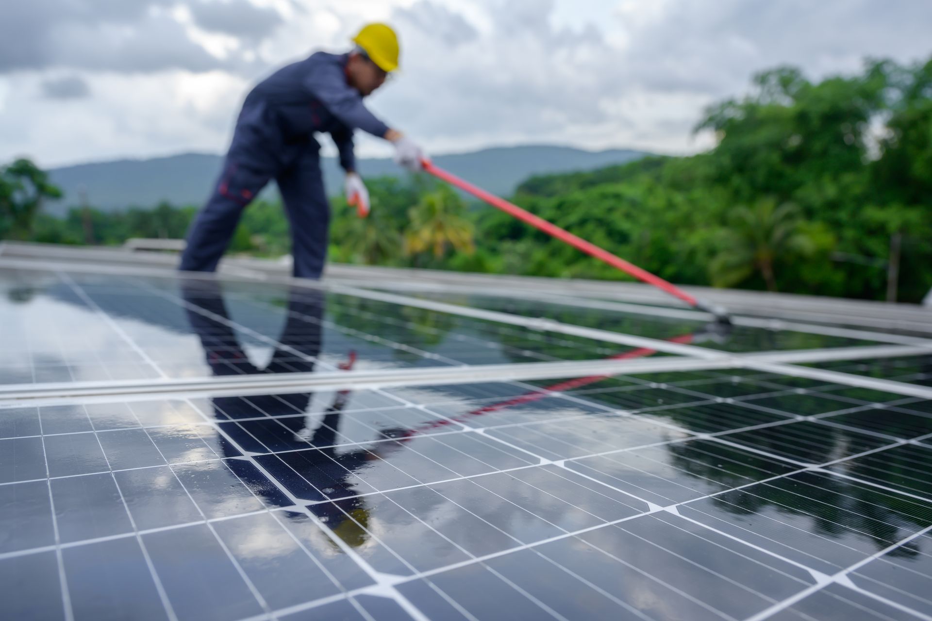 Worker cleaning solar panels on a rooftop with a long pole, wearing a yellow hard hat.