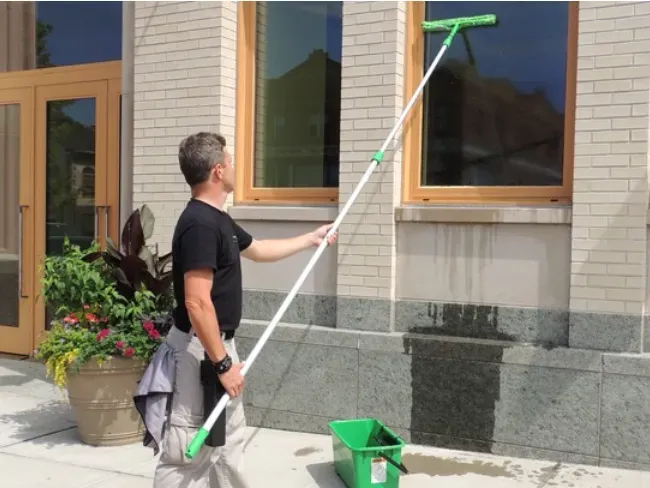 Man using a long-handled window squeegee to clean an exterior window beside a building entrance