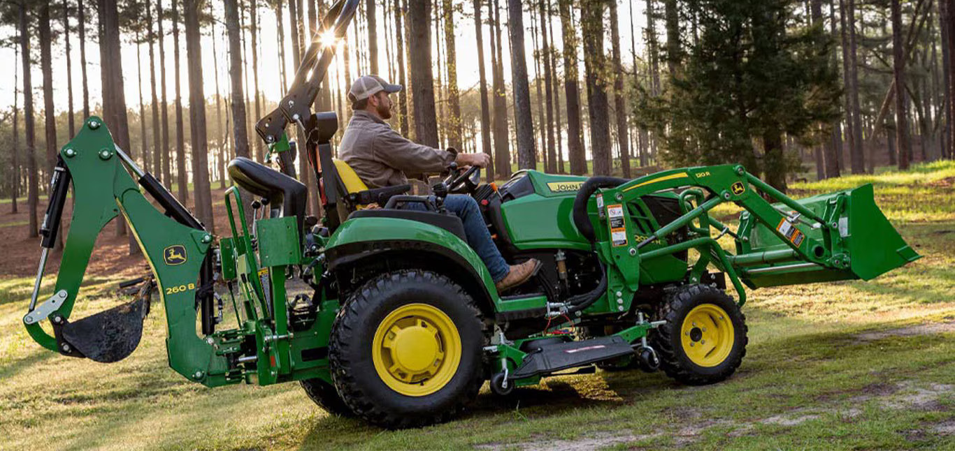 A man driving a green John Deere tractor with a backhoe in a wooded area.