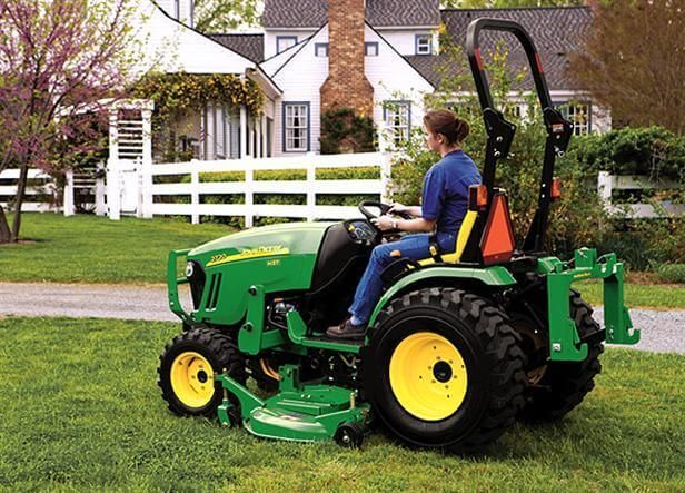 A woman driving a green and yellow John Deere tractor on a lawn in front of a white house.