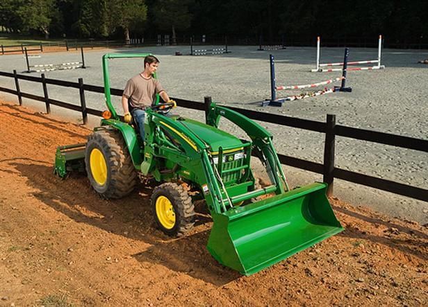 Man driving a green John Deere tractor with a front bucket on a dirt track near a fence.