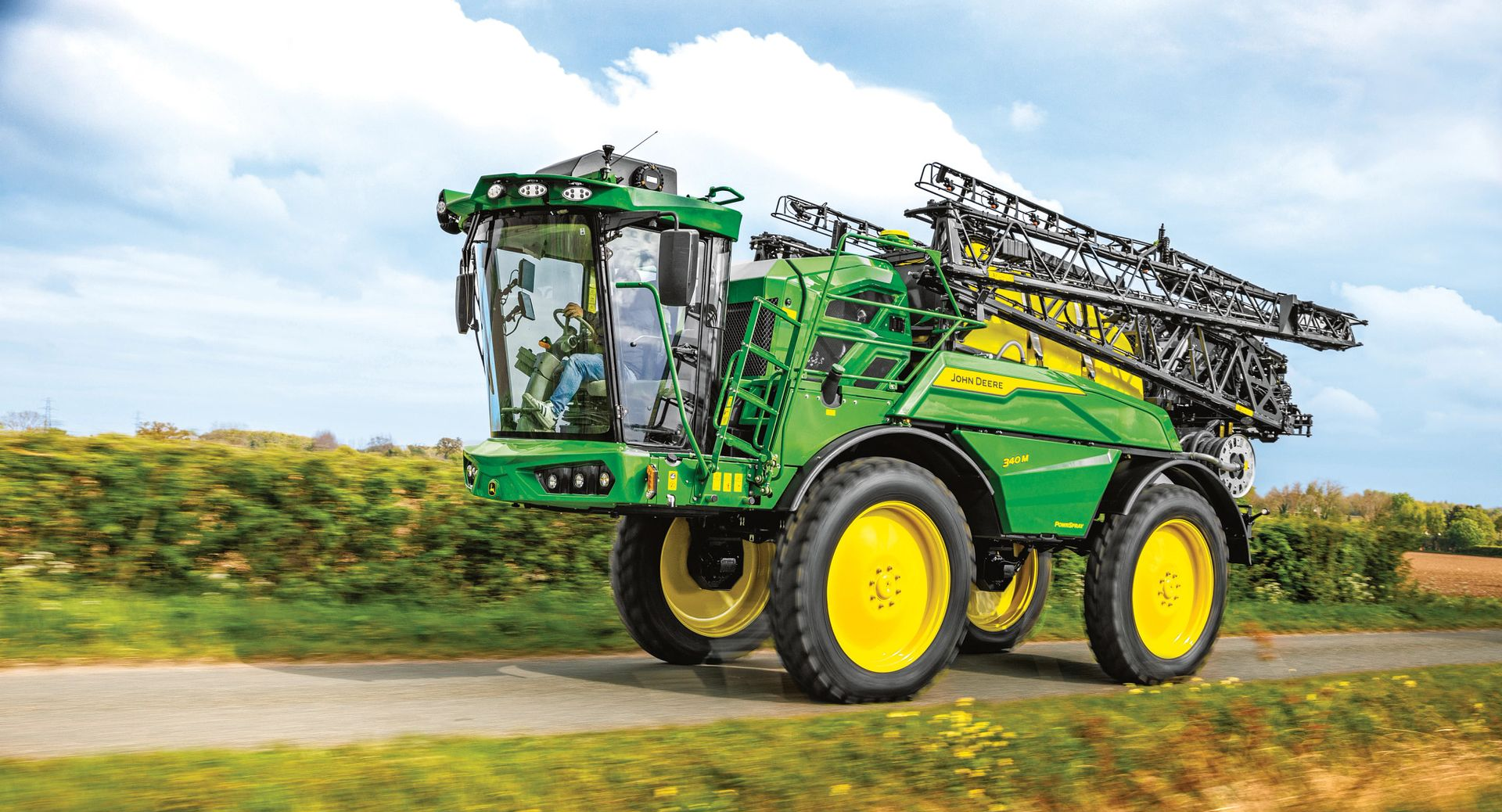 Green John Deere agricultural sprayer driving on a road, booms extended. Yellow wheels.