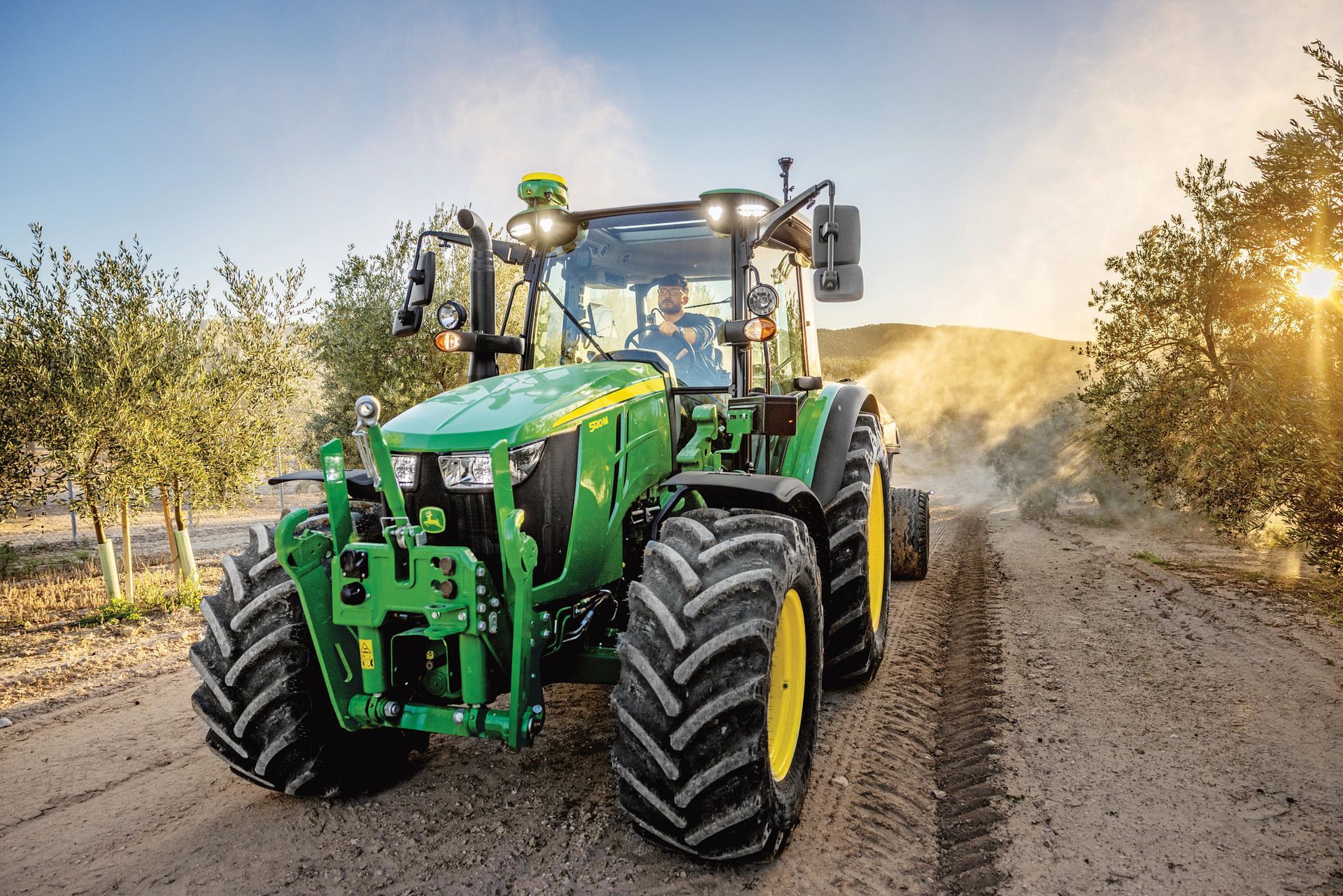 Green John Deere tractor driving on a dirt road with the sun setting.