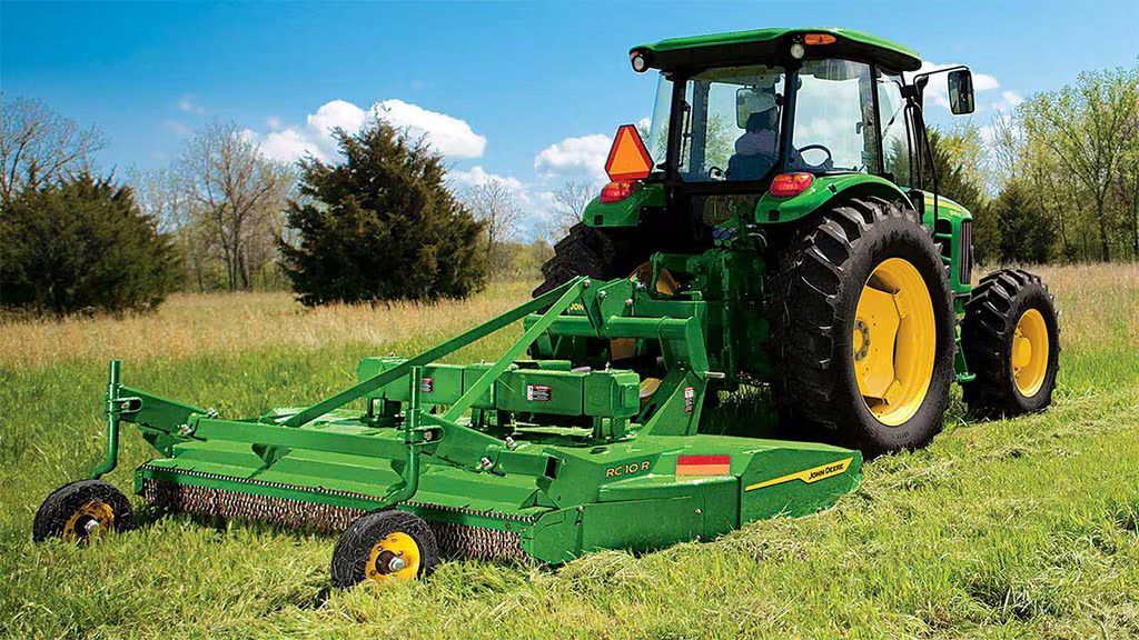 Green John Deere tractor with a mower cutting tall grass in a field. Yellow wheels and a blue sky are visible.