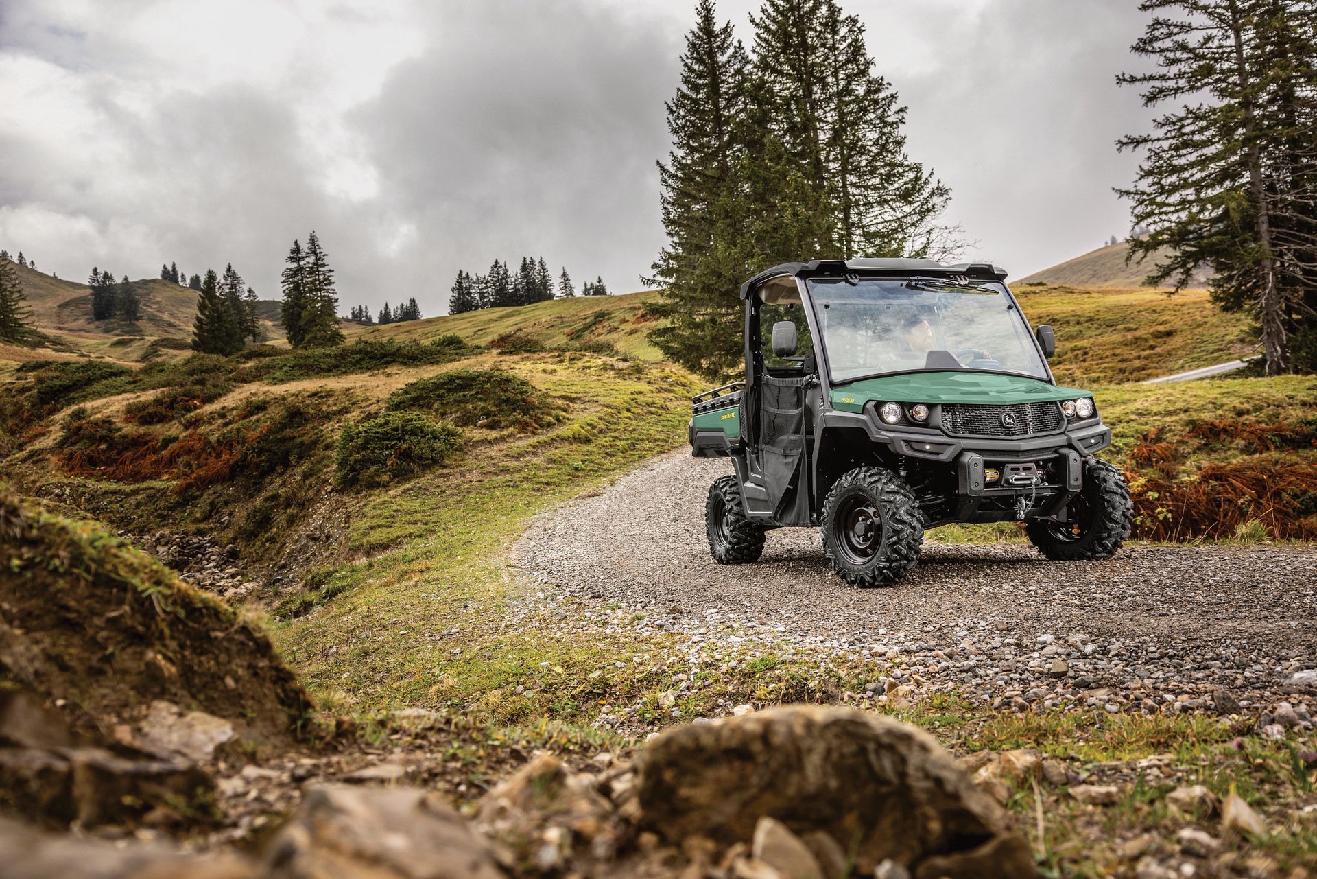 Green utility vehicle on a gravel path in a mountainous, wooded area under a cloudy sky.
