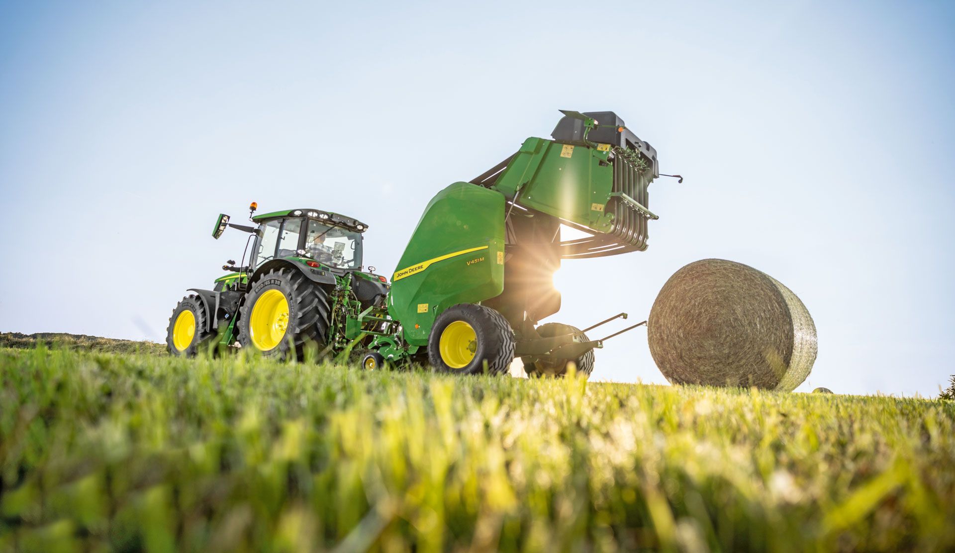 A green John Deere tractor baling hay in a sunny field. A large, round bale is being ejected.