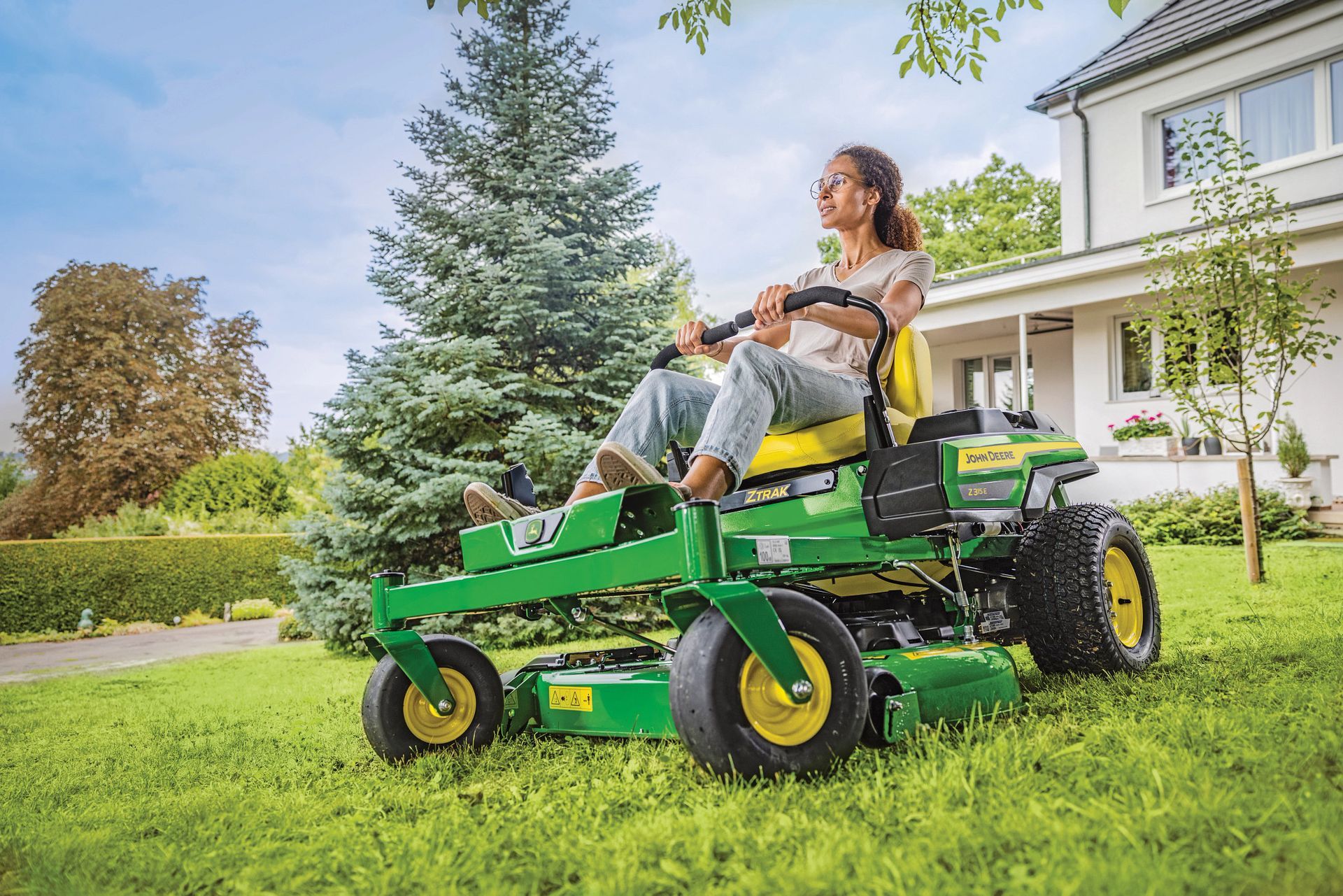 Woman on a green John Deere riding lawn mower, cutting grass in front of a house on a sunny day.