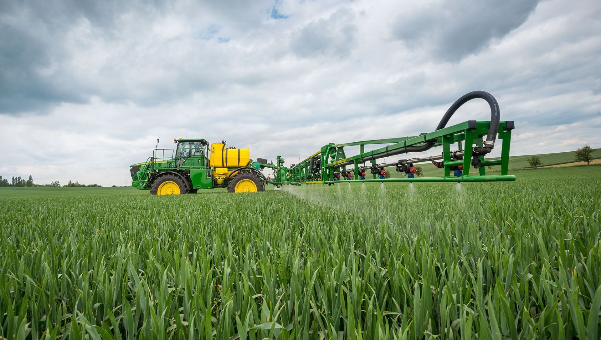 John Deere sprayer in a green field, spraying crops under a cloudy sky.
