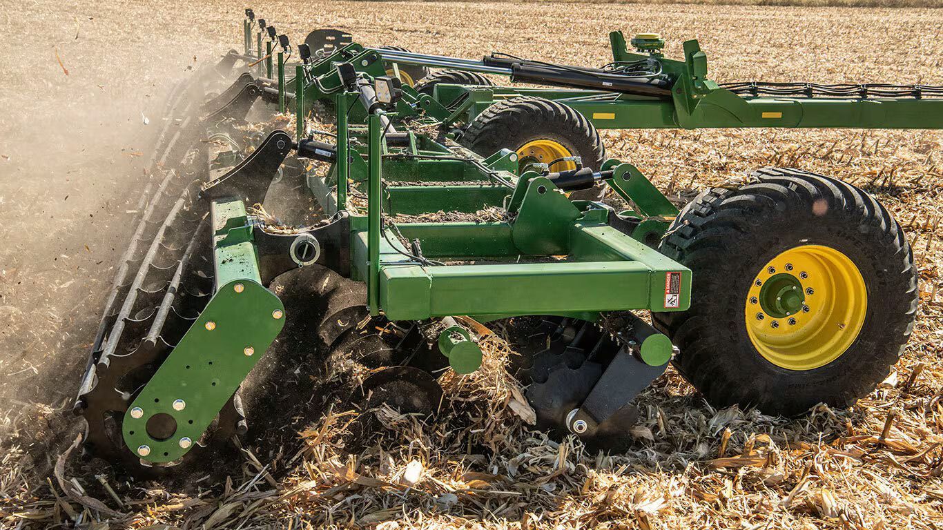 Green John Deere farm implement tilling a field, kicking up soil and debris. Yellow wheels and a sunny agricultural setting.