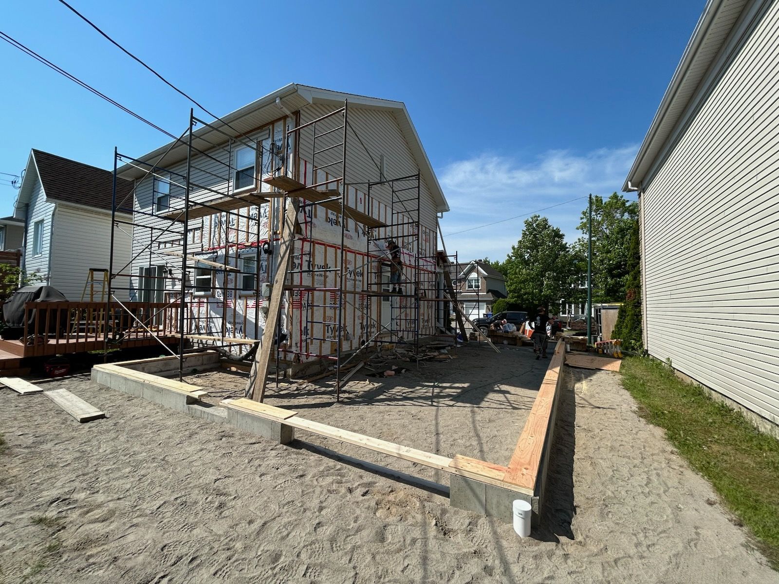 Maison en construction, échafaudages contre le bardage, fondations en bois et cour en gravier.