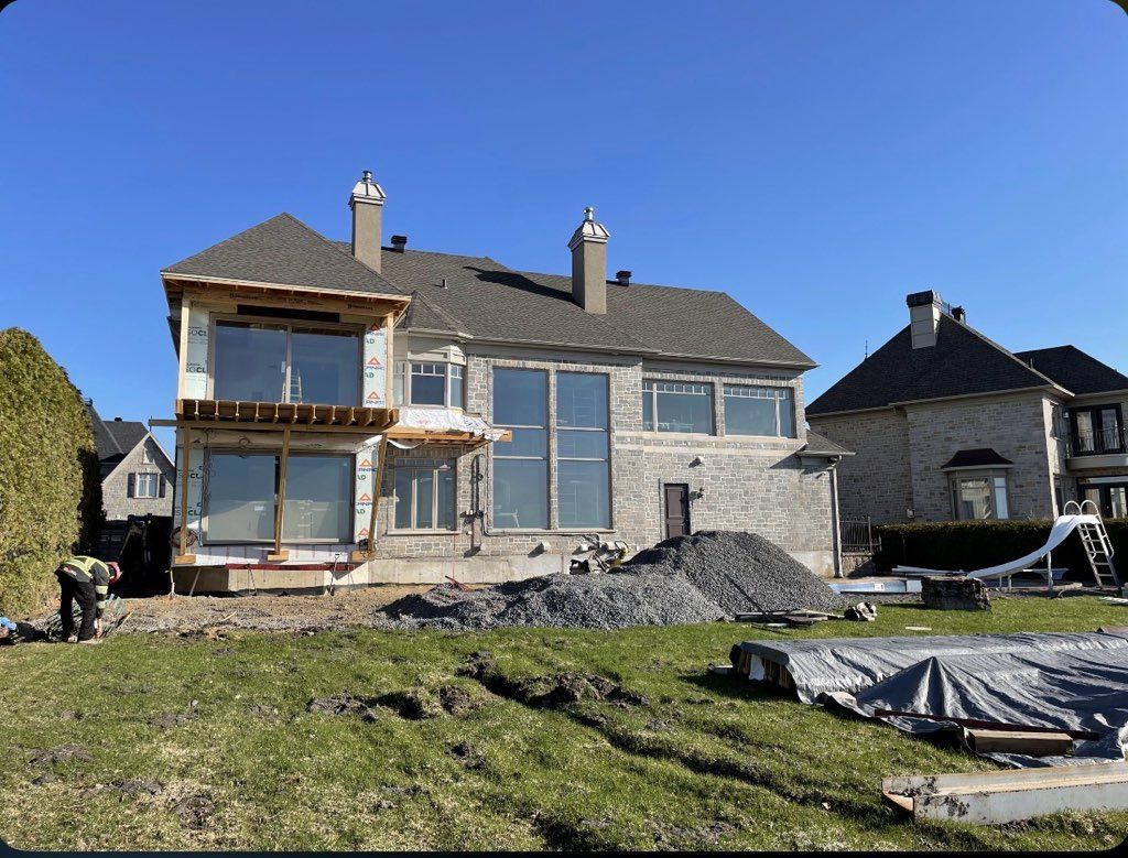 Maison en construction, vue extérieure. Façade en pierre, grandes fenêtres, tas de gravier devant.