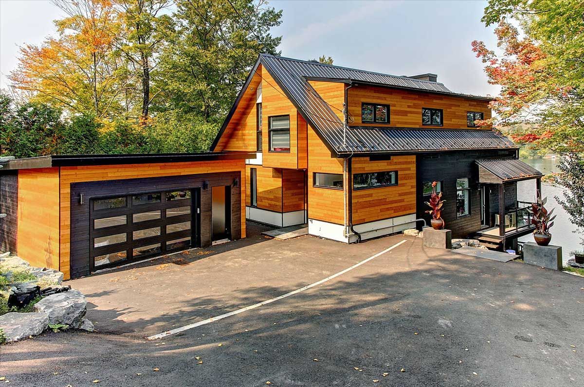 Maison moderne revêtue de bois avec garage attenant, toit foncé et allée pavée, entourée d'arbres.