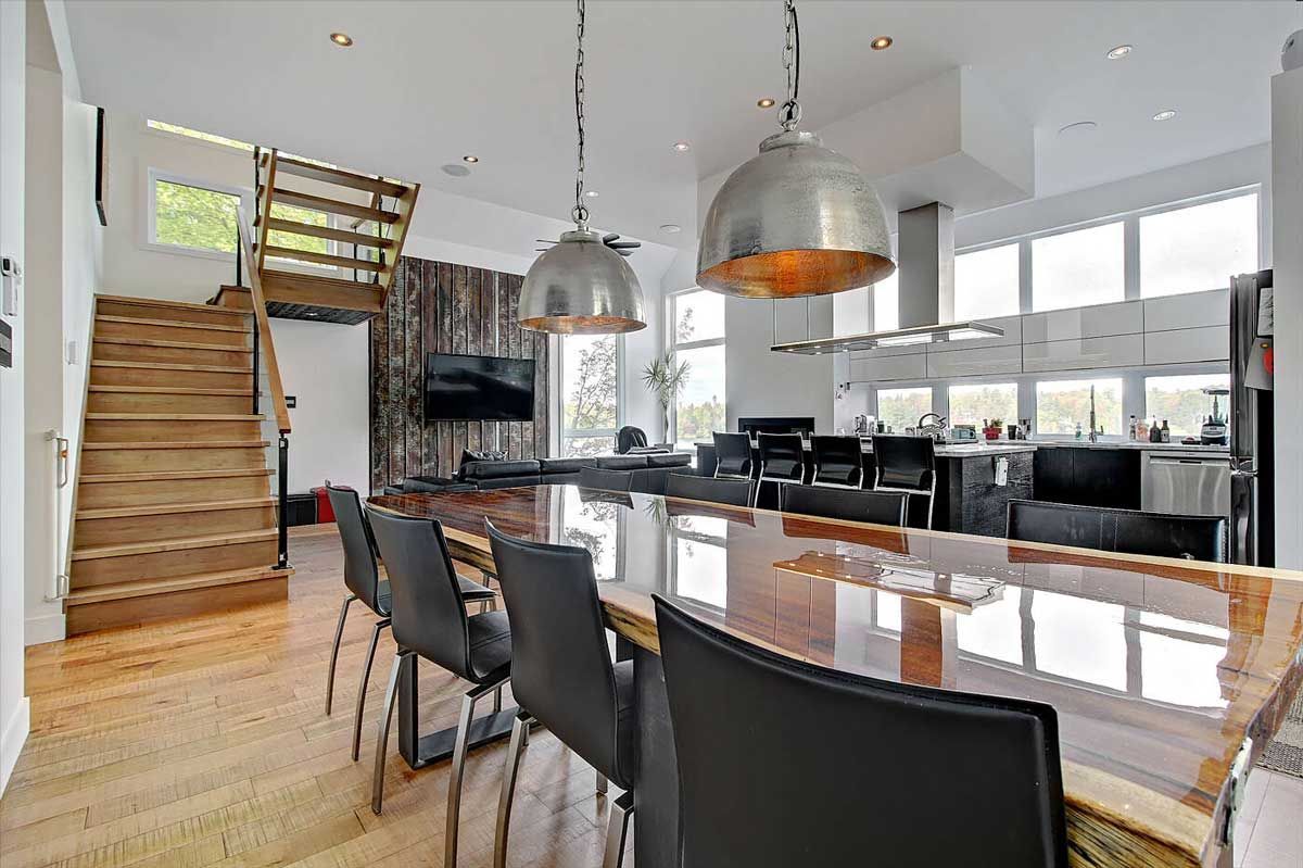 Cuisine moderne avec escalier en bois, îlot central avec sièges, luminaires en acier inoxydable et grandes fenêtres.