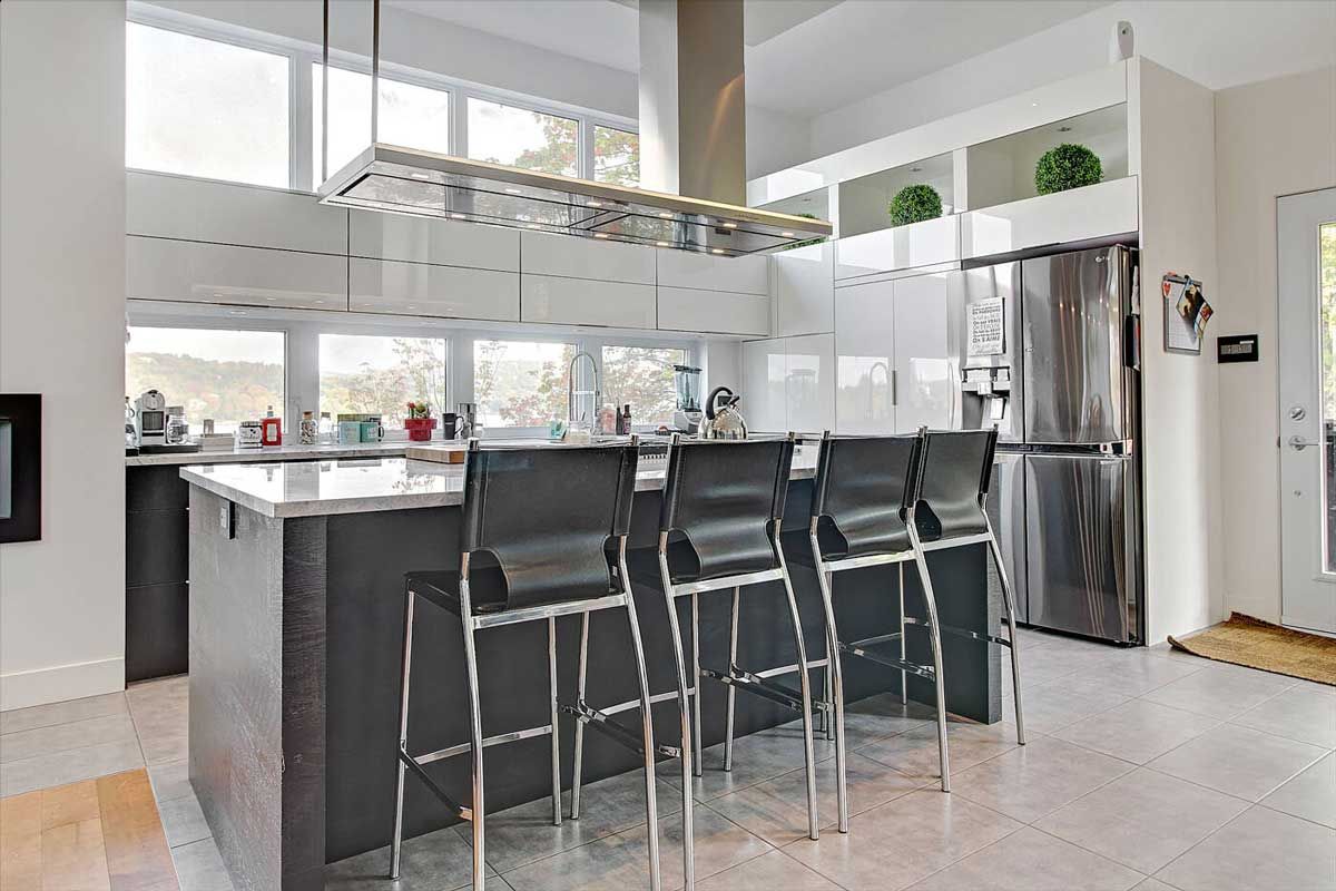 Cuisine moderne avec îlot gris, tabourets de bar noirs, appareils électroménagers en acier inoxydable et grandes fenêtres.