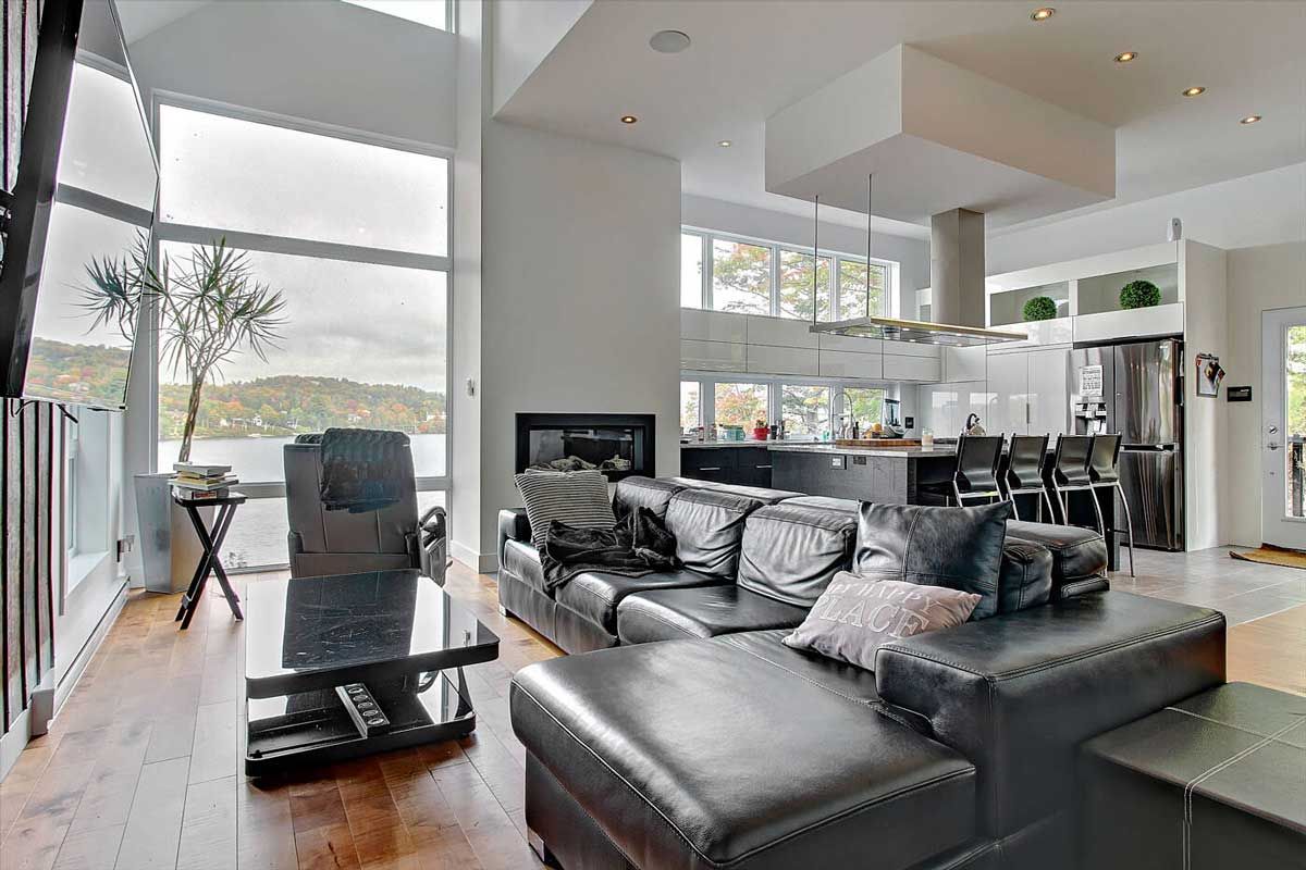 Salon avec de grandes fenêtres donnant sur un lac. Canapé en cuir noir, cheminée et cuisine visibles.