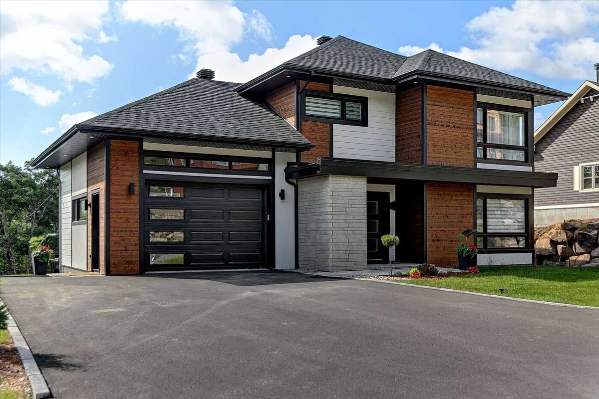 Maison moderne à deux étages avec boiseries noires, briques et bardage blanc. Porte de garage noire et allée asphaltée.