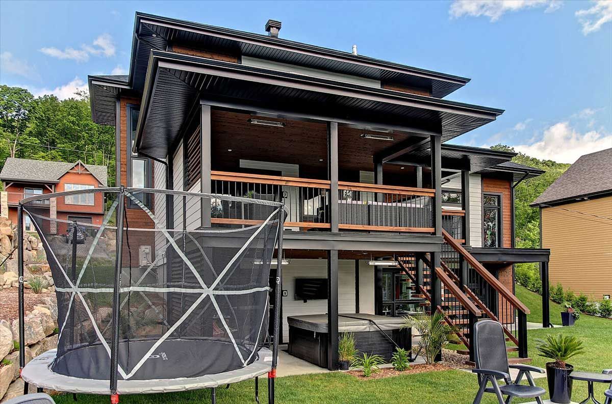 Maison à deux étages avec des boiseries noires et une terrasse ; trampoline dans le jardin.