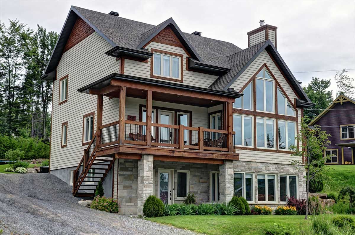 Maison à deux étages avec porche en bois et hautes fenêtres ; allée et pelouse à l'avant, arbres à l'arrière-plan.