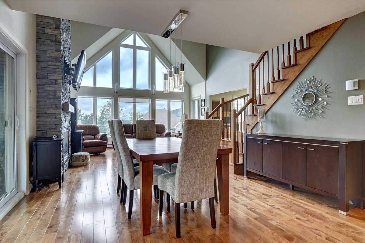 Salle à manger avec table et chaises en bois, attenante au salon avec grandes fenêtres, cheminée et escalier.