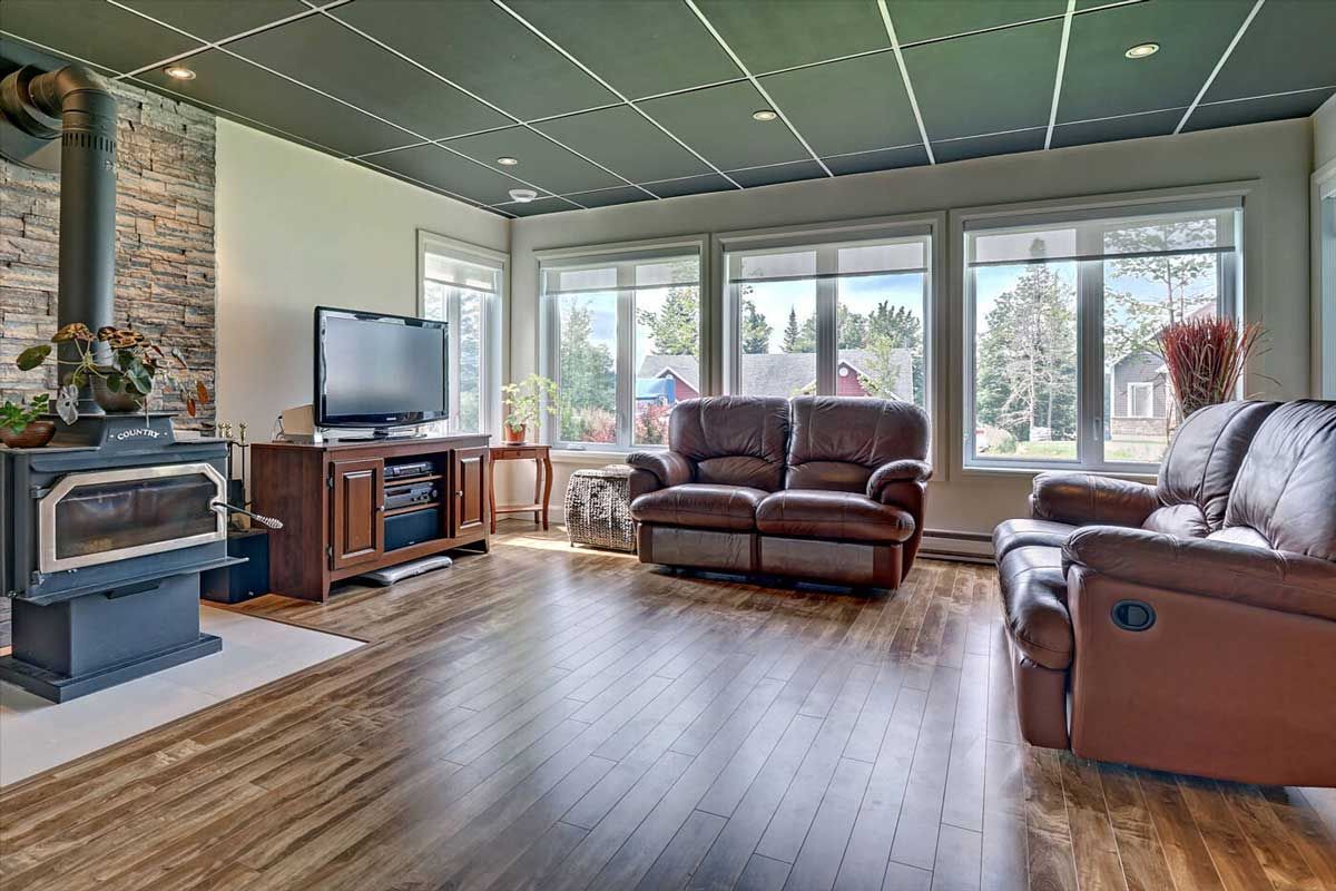 Salon avec parquet, cheminée en pierre, poêle noir, canapés en cuir marron et grandes fenêtres.