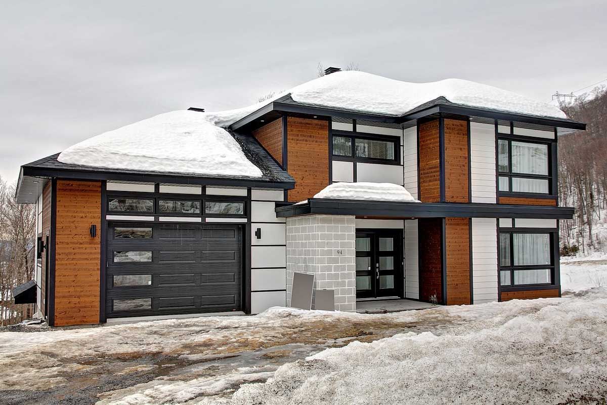 Maison moderne avec boiseries noires, bardage en bois et toit enneigé. Garage à gauche.
