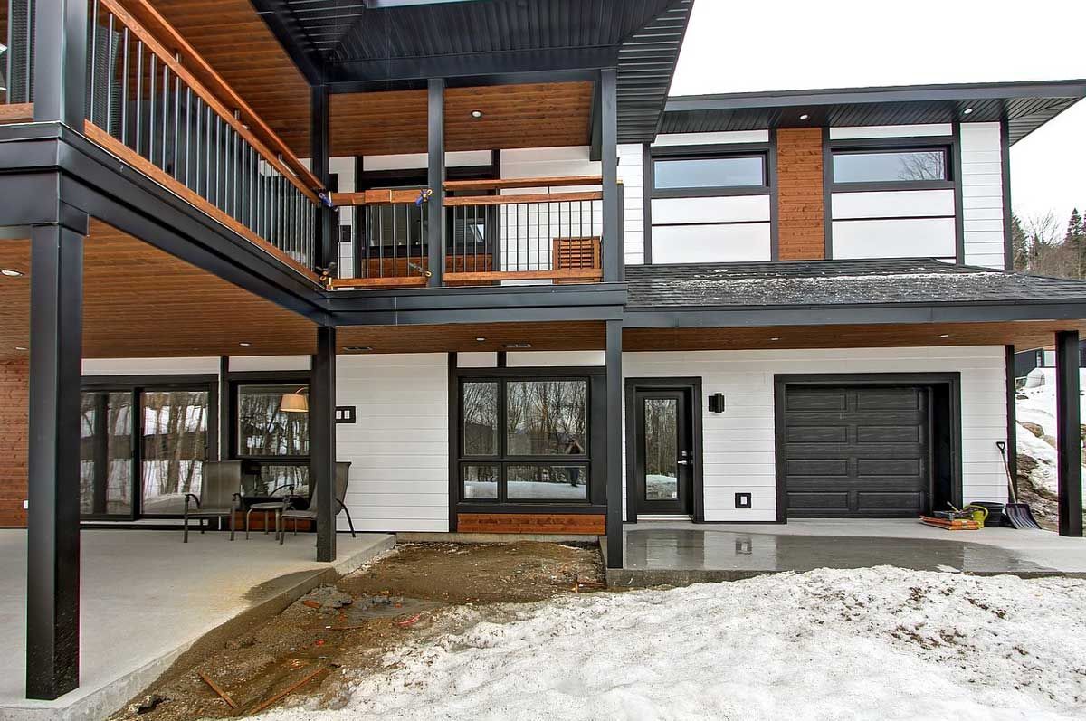Maison moderne à deux étages avec des finitions noires, des éléments en bois et une terrasse couverte dans un paysage enneigé.