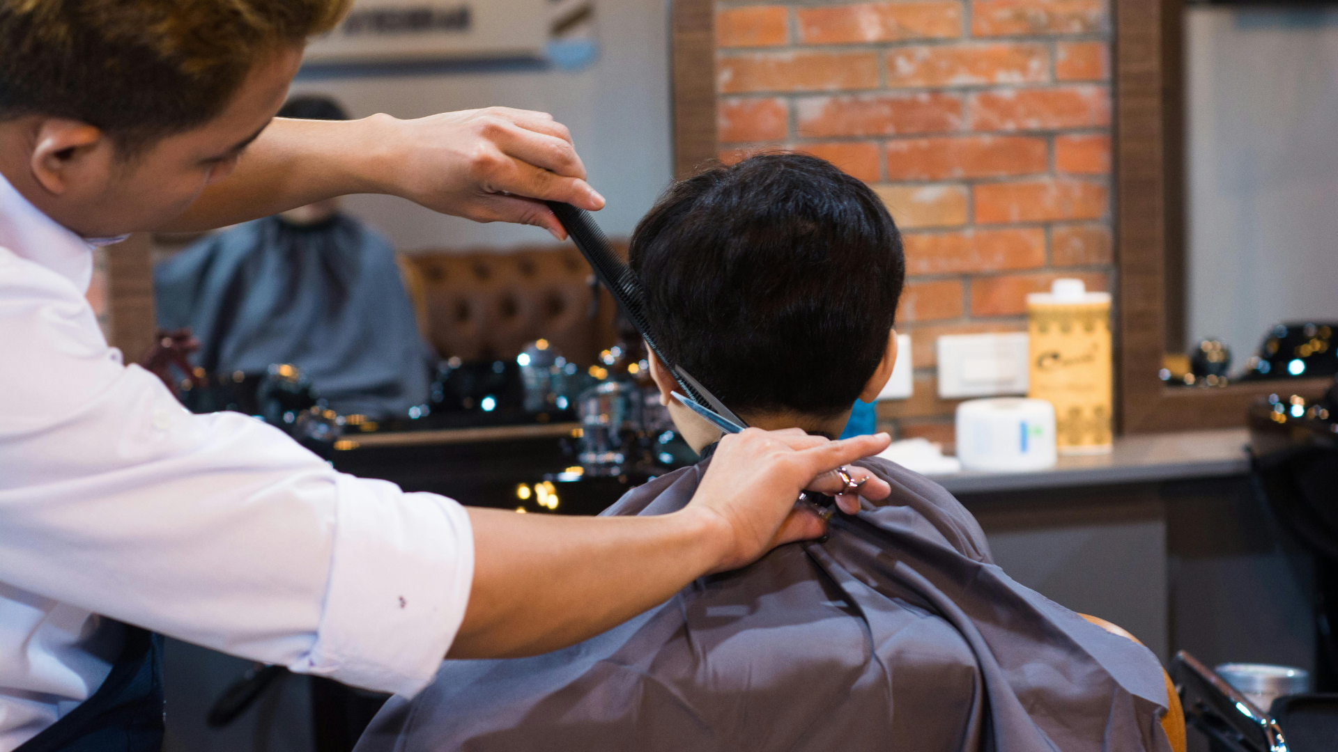 A barber using scissors and a comb to trim a person's hair in a salon with a brick-patterned wall in the background.