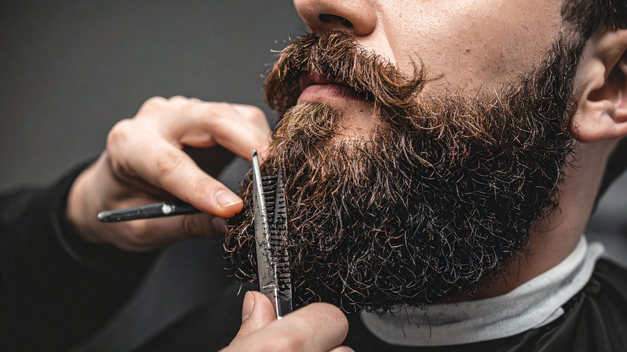 A barber using scissors and a comb to trim and shape a client's thick, dark, groomed beard.