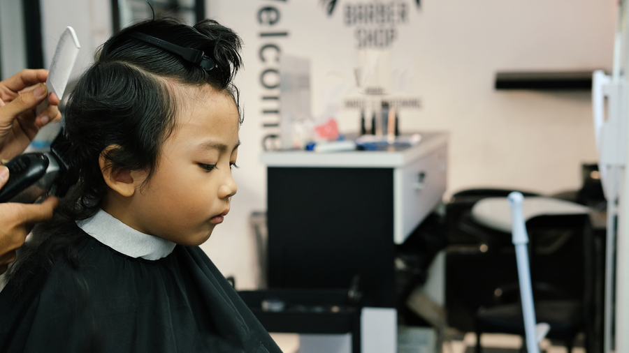 A barber cuts the hair of a person wearing a black cape in a barbershop.