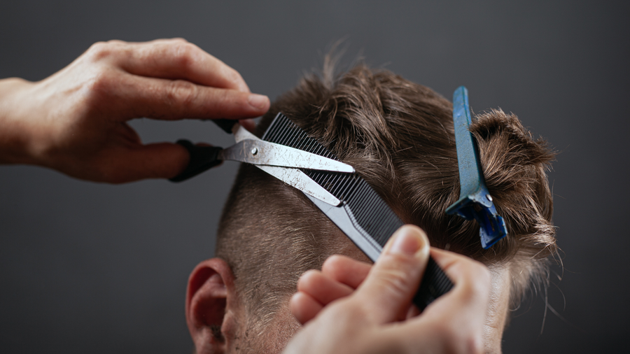 A hairstylist uses scissors and a black comb to trim a person's hair, with a blue sectioning clip holding the hair back.