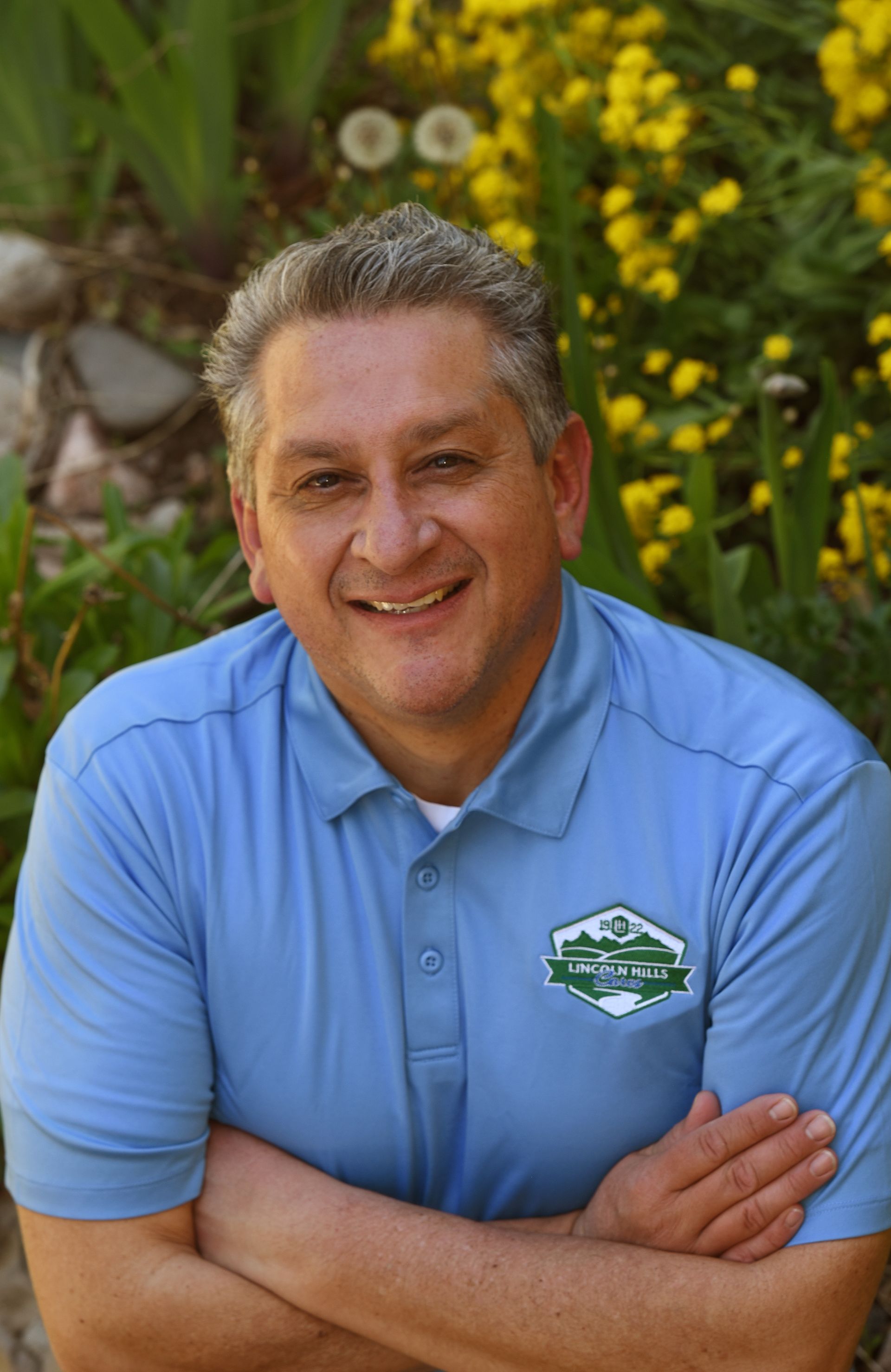 A man in a blue shirt is standing in front of a garden with his arms crossed.