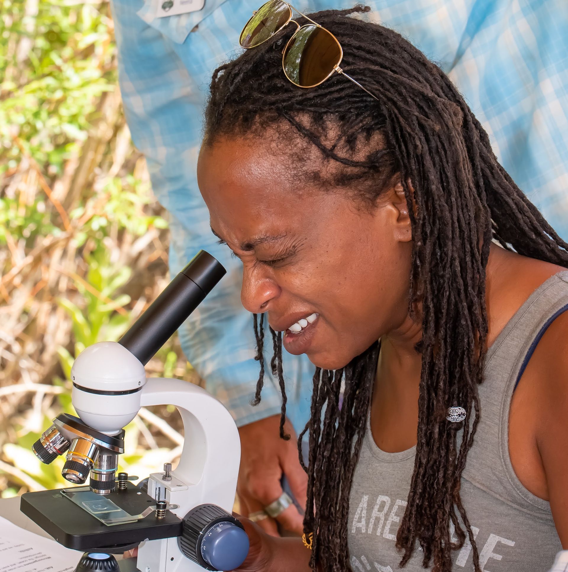 A woman with dreadlocks looks through a microscope while wearing sunglasses