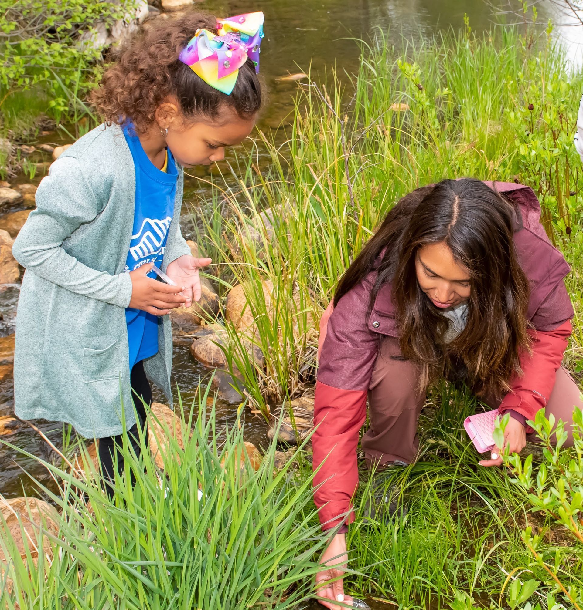Two women are kneeling in the grass near a river.