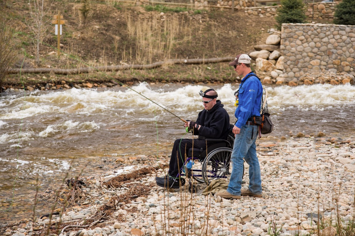 A man in a wheelchair is fishing with a man standing next to him.