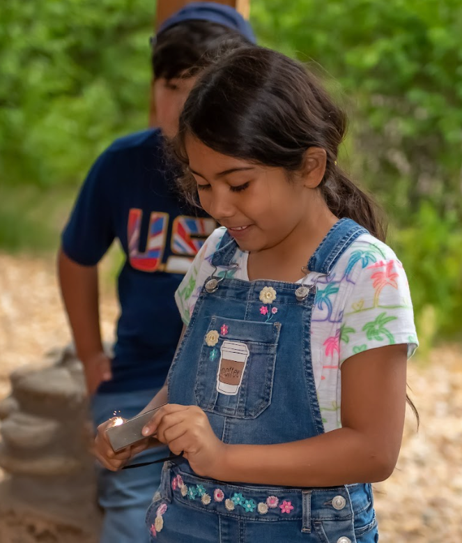 A young girl in overalls is looking at a cell phone.