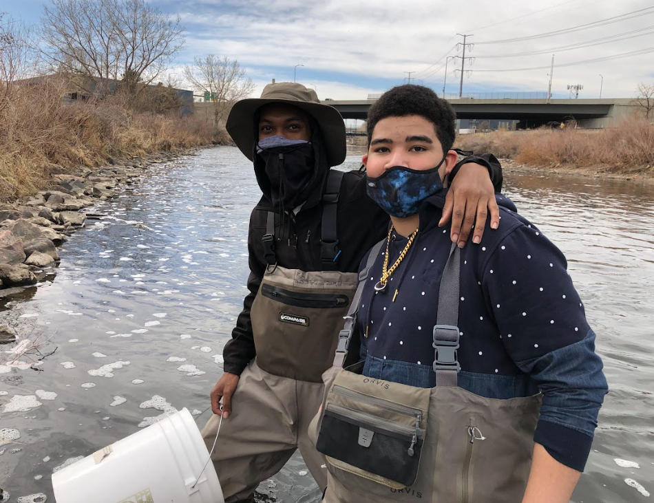 Two young men wearing masks are standing next to a river.
