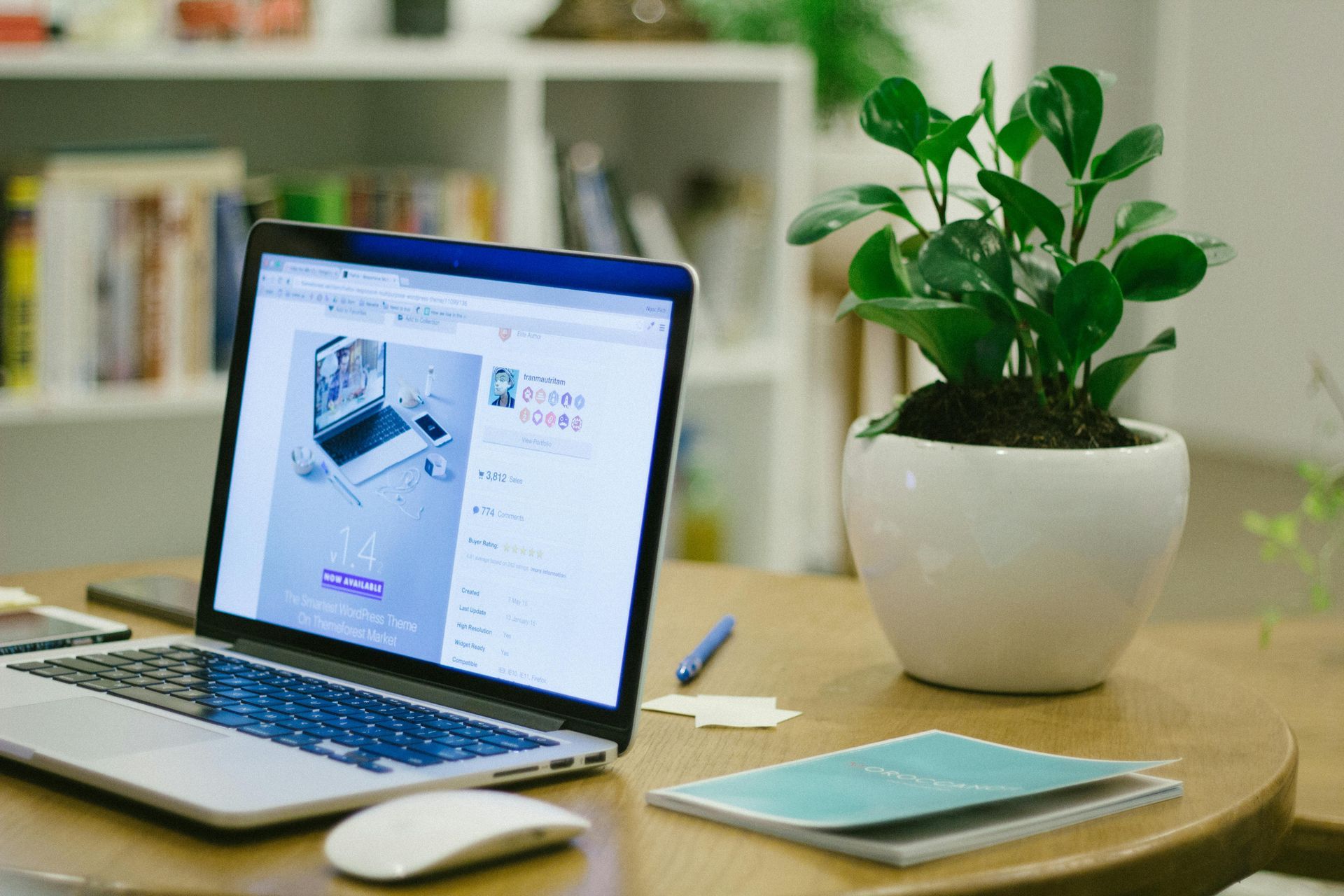 Laptop on a wooden table with a plant, mouse, notepad, and pen. White bookshelf in the background.