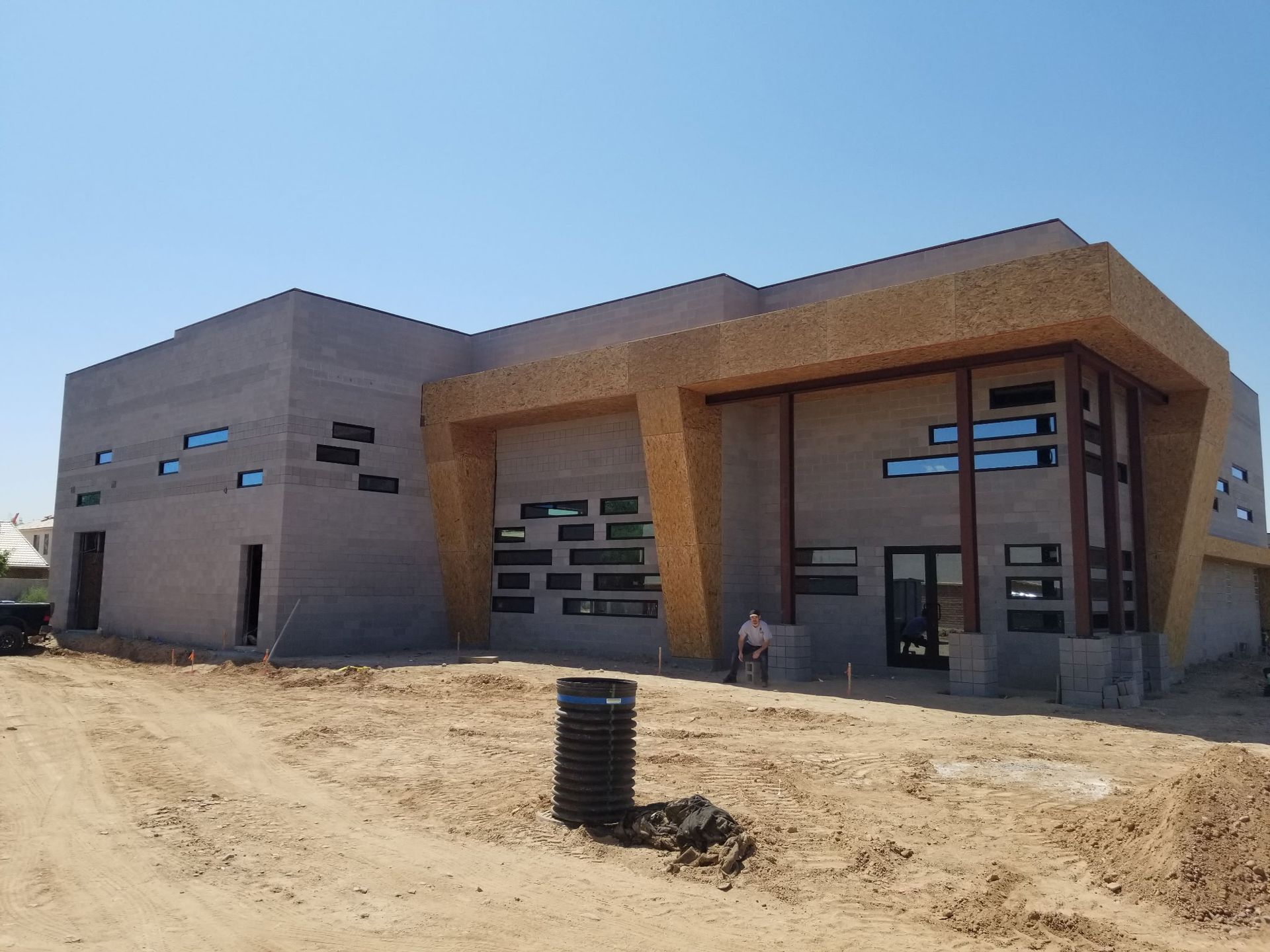 Modern building under construction, tan and gray concrete exterior, numerous windows, open dirt lot.