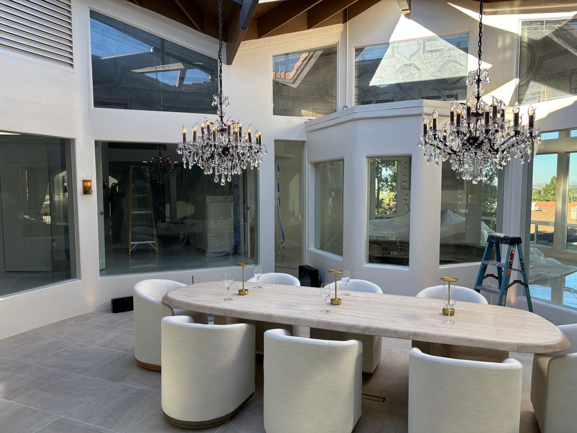 A dining room with a long stone table, cream chairs, and two ornate crystal chandeliers under a white vaulted ceiling.