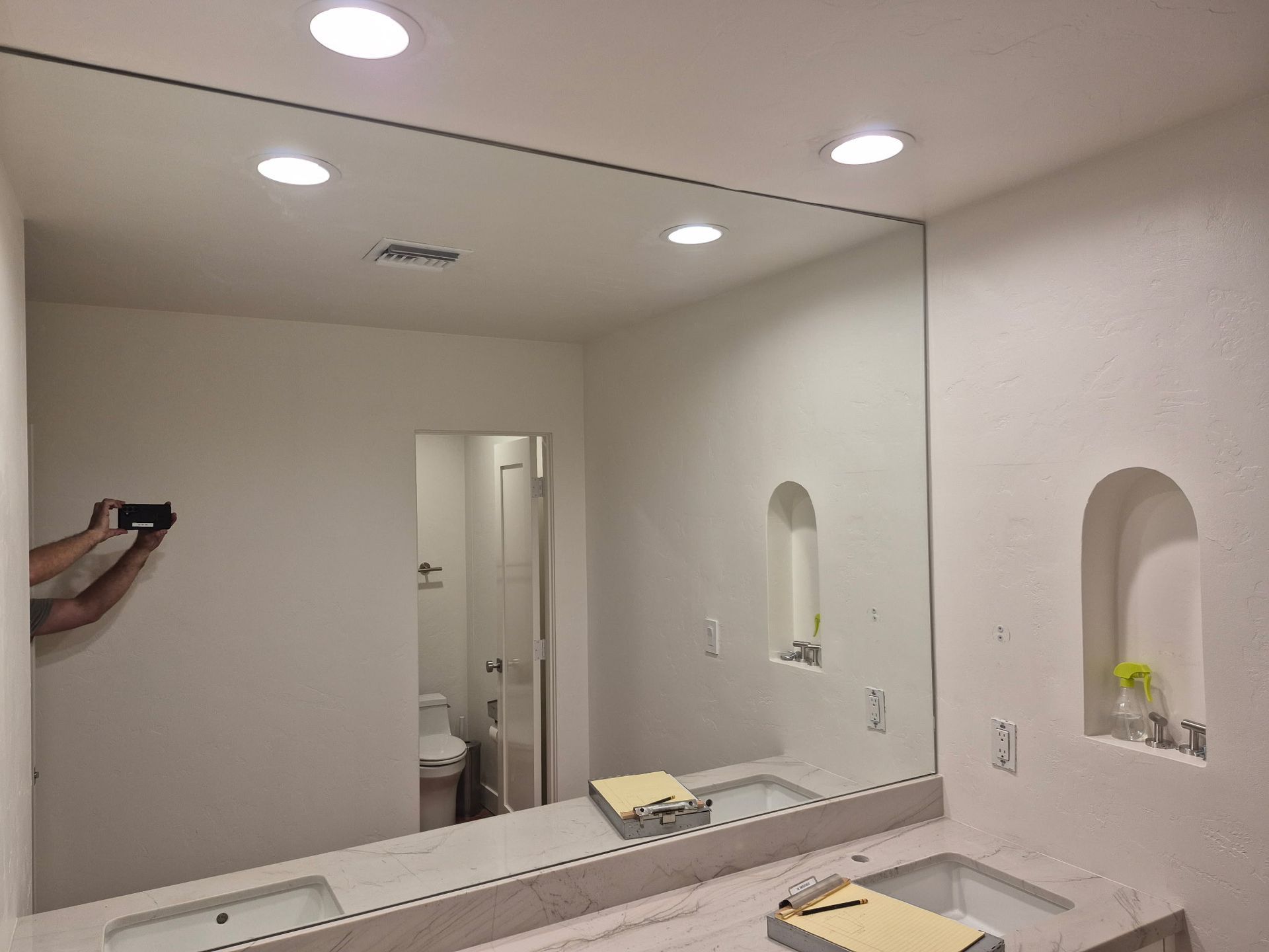 Bathroom with large mirror above double sinks, recessed lighting, and a person taking a photo.