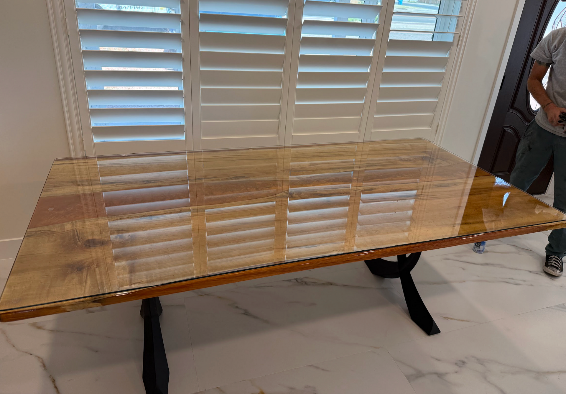 A glossy wooden table with black legs is in front of a window with blinds, person at the right.