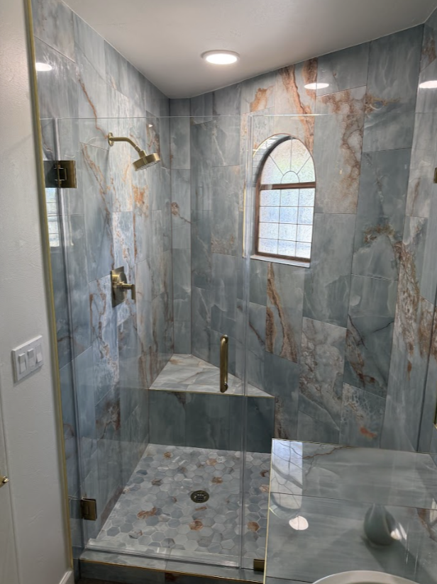 Blue marbled tile shower with glass doors, gold fixtures, and arched window.