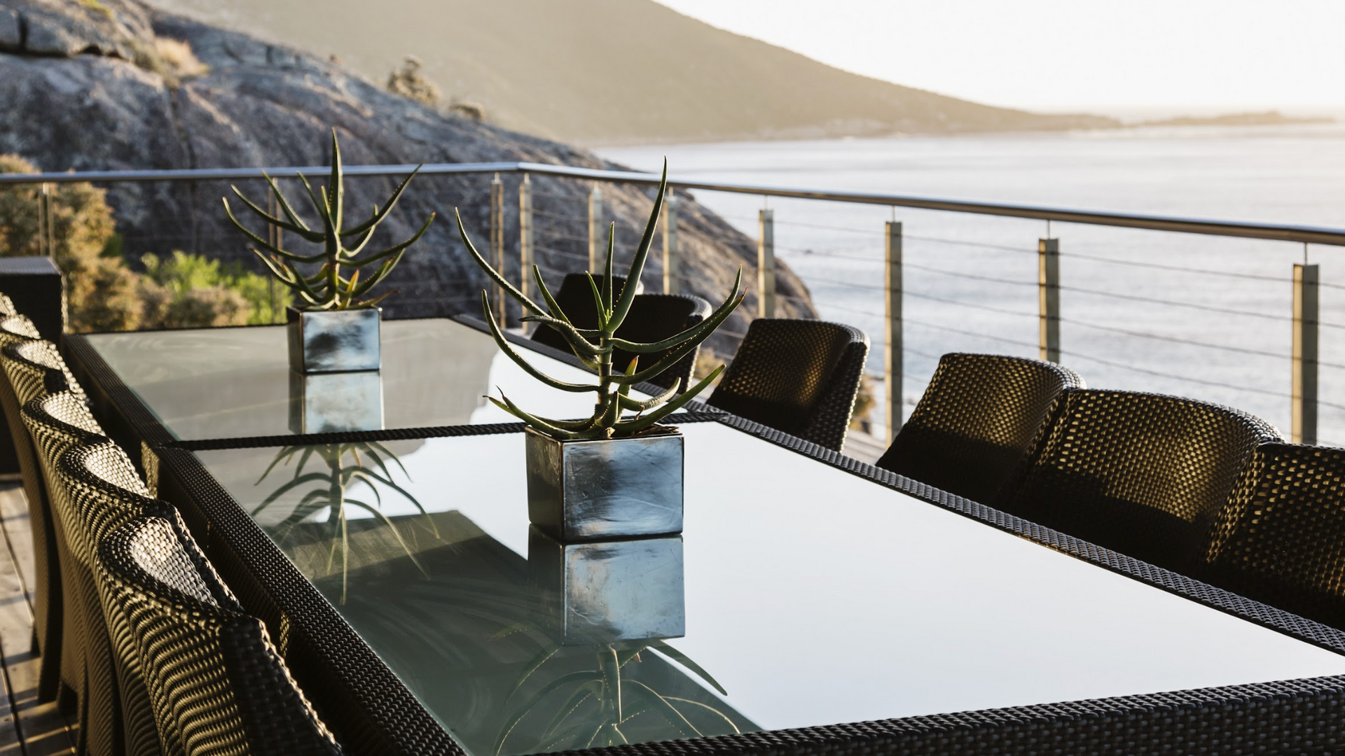 Outdoor dining table with coastal views, dark wicker chairs, glass top, and potted plants.