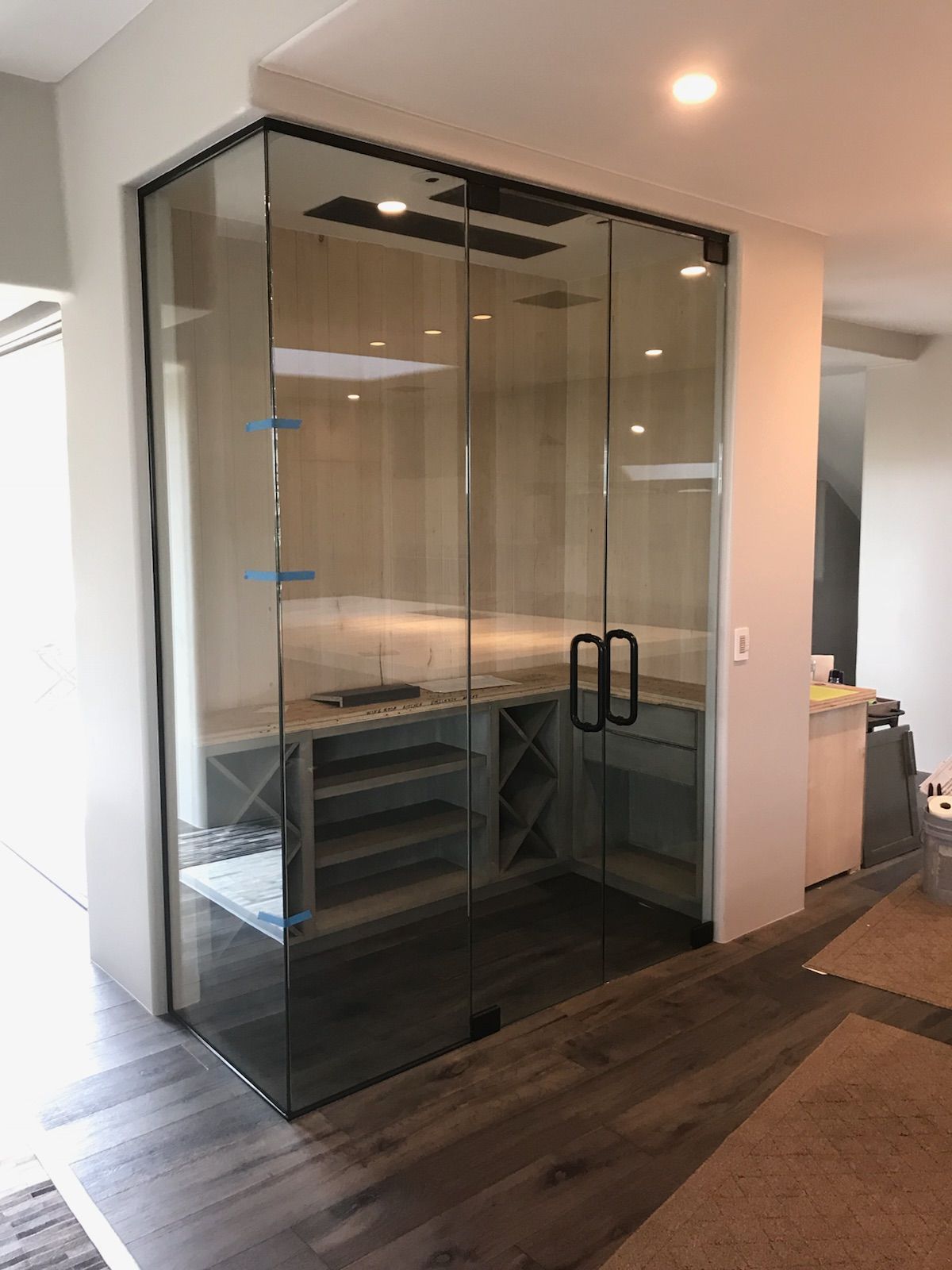 Glass-enclosed wine cellar with wooden shelves and dark door handles, built into a room with wood floors.