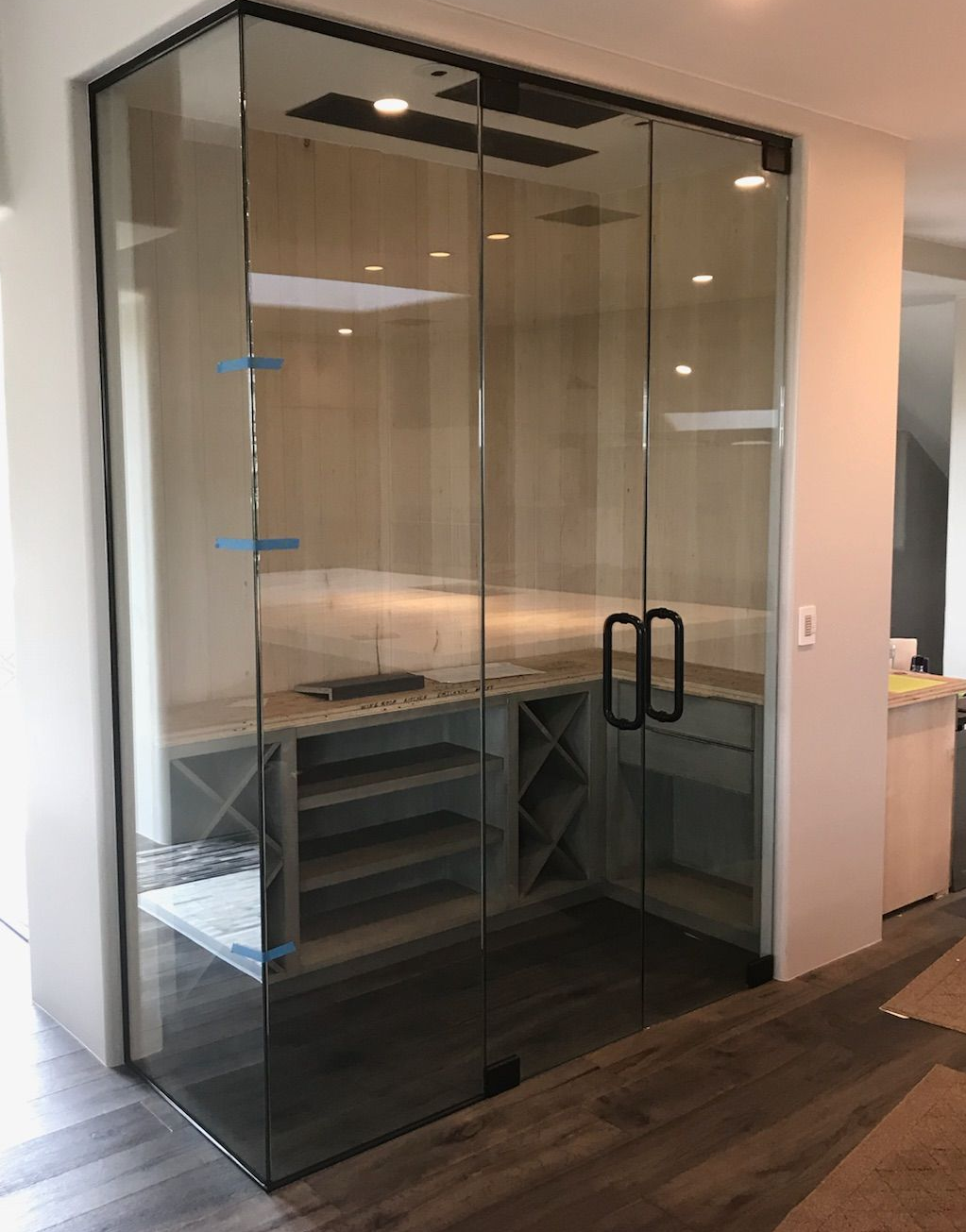 Glass-enclosed wine cellar with black frame and handle. Interior features wooden shelves, gray cabinets, and wine racks.