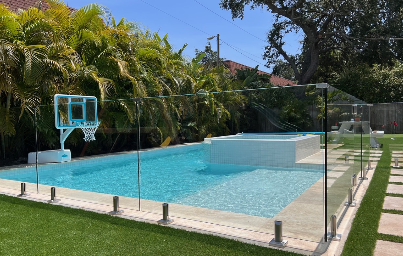 Pool with clear glass fencing and a basketball hoop, surrounded by greenery.