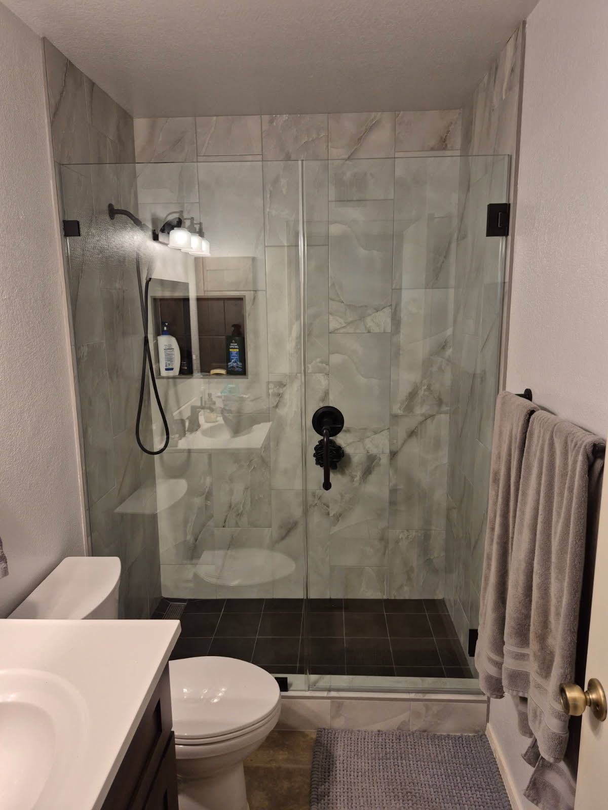 Small bathroom with glass shower, toilet, sink, towel, and gray tile walls