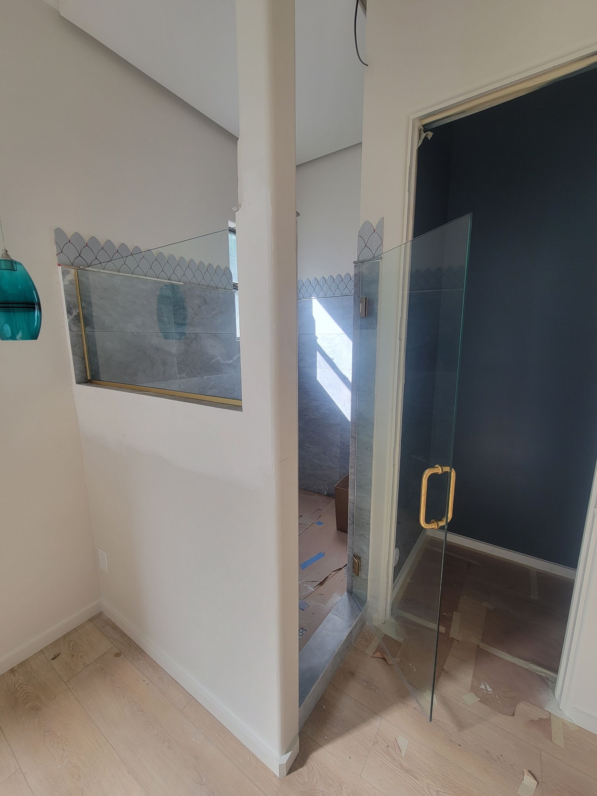 Bathroom with clear glass shower door and wall. White walls, wood floor, and gold handle.