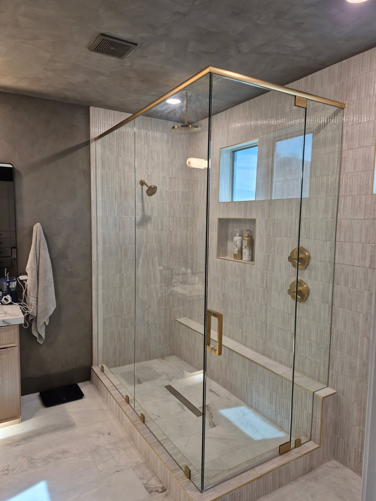 Modern glass shower with gold trim, stone walls, and a bench.